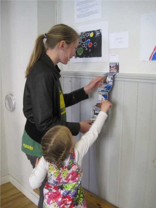 Barn og voksen studerer en remse med bilder som henger på en vegg.