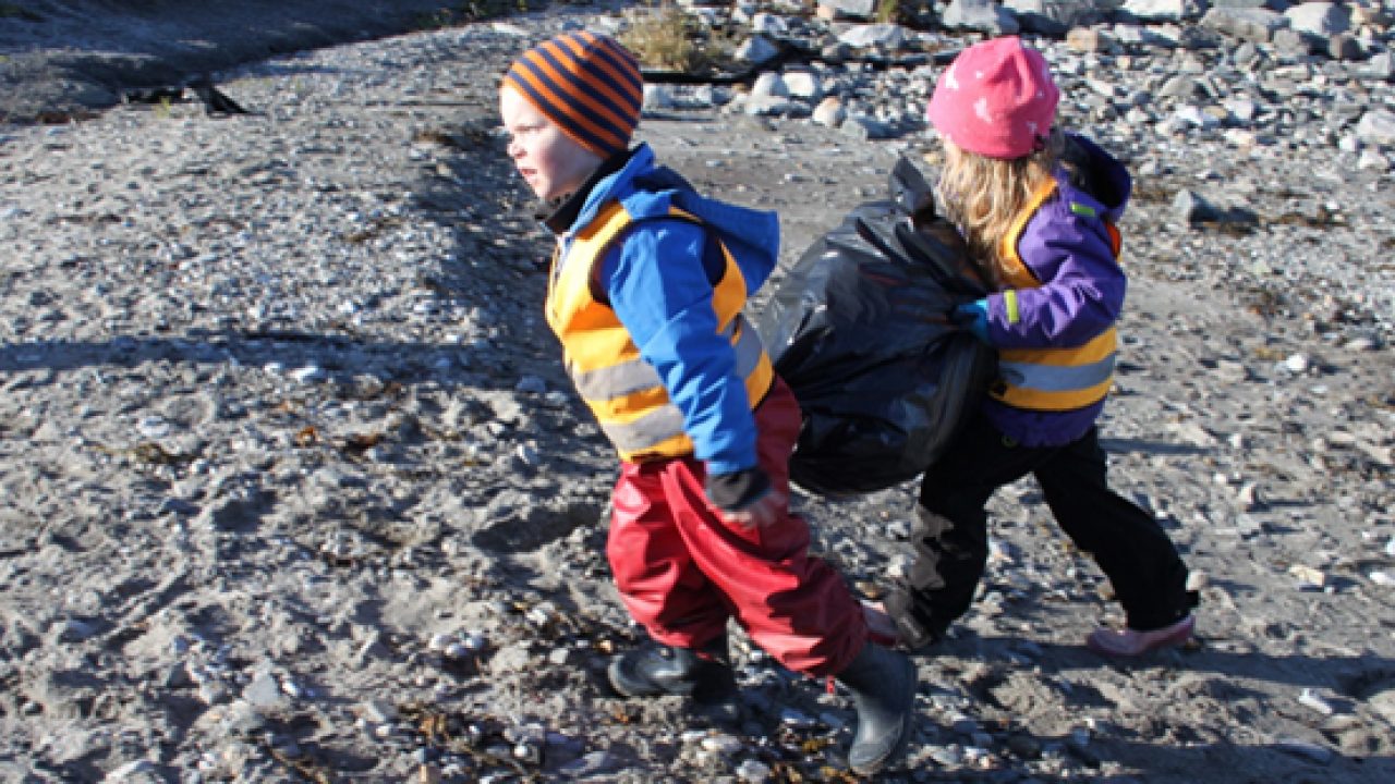 Barn i fjæra bærer svart søppelsekk mellom seg