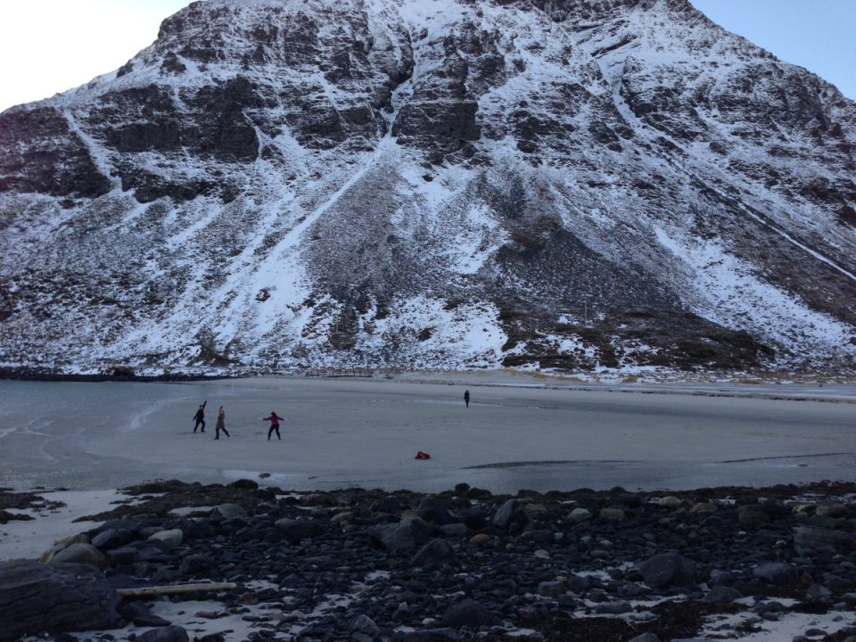 Naturfoto. En liten gruppe mennesker danser på en strand rett ved et snødekt fjell.