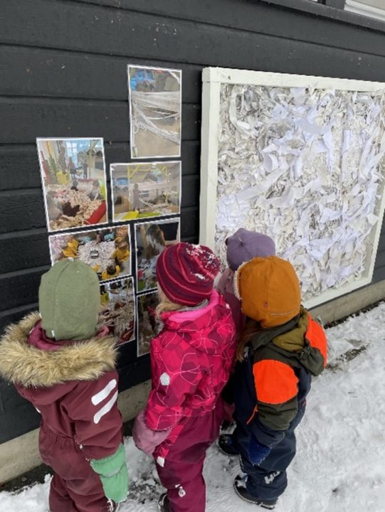 Barn står ute og ser på en vegg med bilder. Bildene er av dem selv og andre barn i barnehagen som utforsker papir.