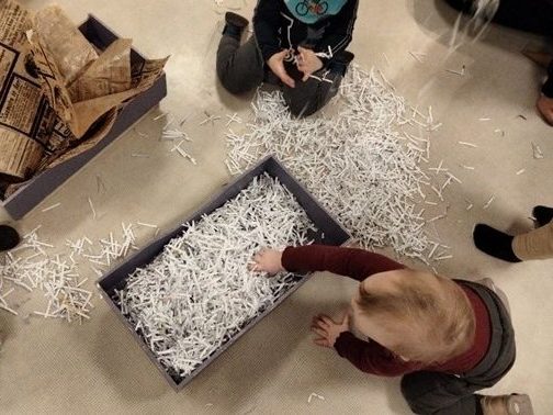 To små barn sitter på gulvet ig utforsker en balje med papirstrimler.