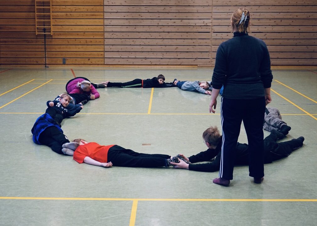 Elever ligger på gulvet i en gymsal og lager sammen en sirkelform. En lærer står like ved.
