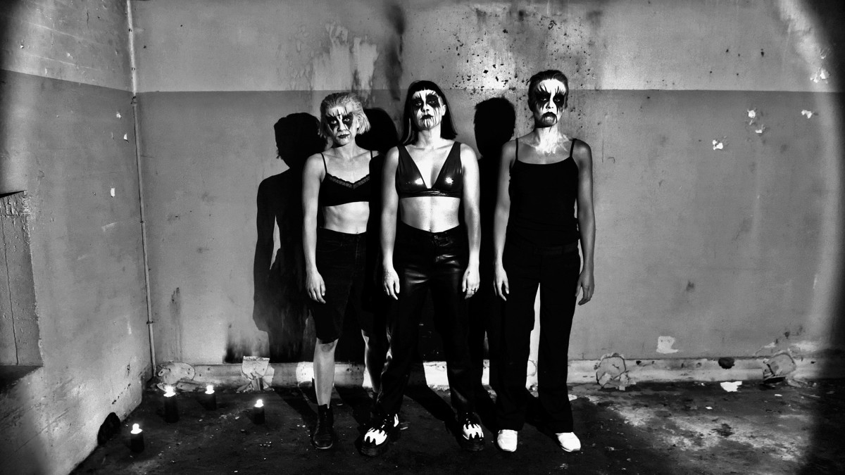 Bilde i sort hvitt. Tre damer står og ser rett i kamera. De er malt sort rundt øynene.
