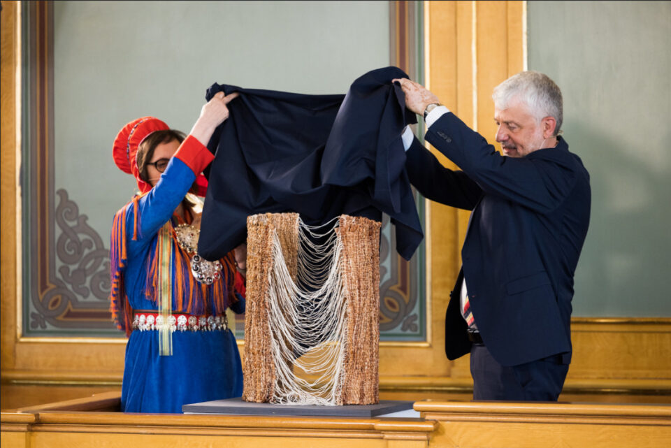 Kvinne i samisk drakt og mann i mørk dress avduker et skulpturalt kunstverk.