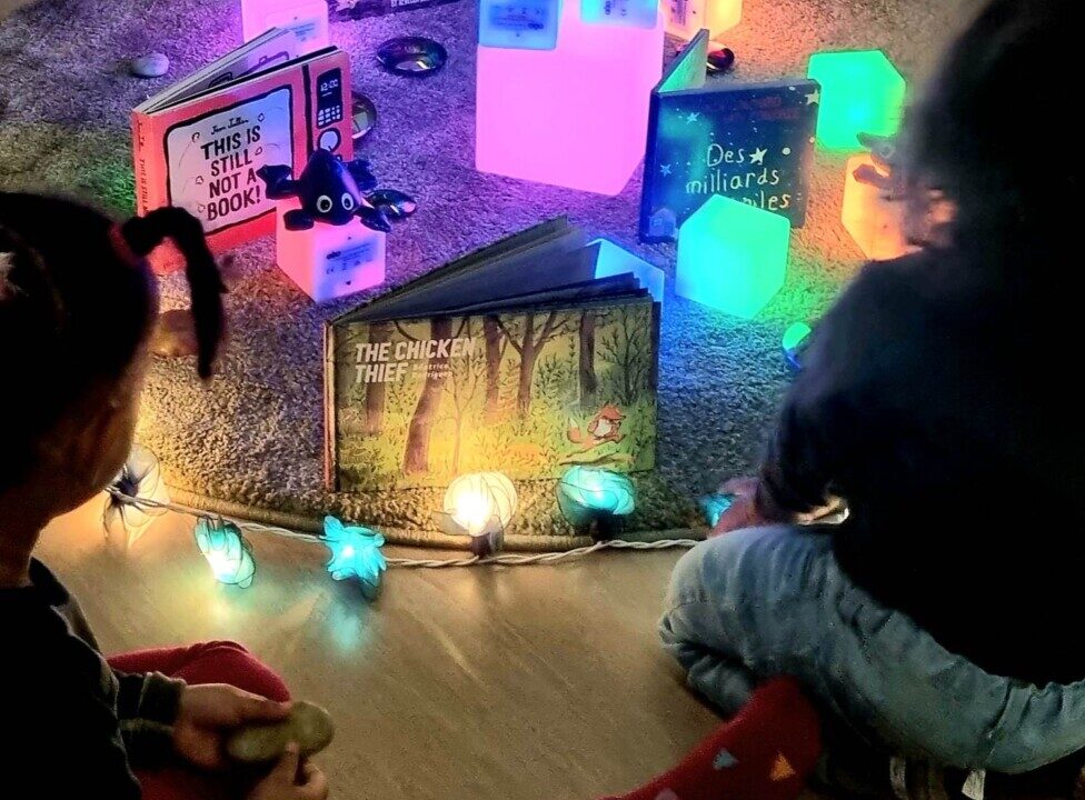 Bøker ligger presentert på et teppe på gulvet. Lyslenker og lysende kuber, og noen leker ligger ved. to barn sitter og esr på bøkene.