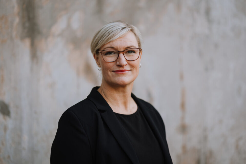 Dame med lyst hår og briller. Svart jakke.